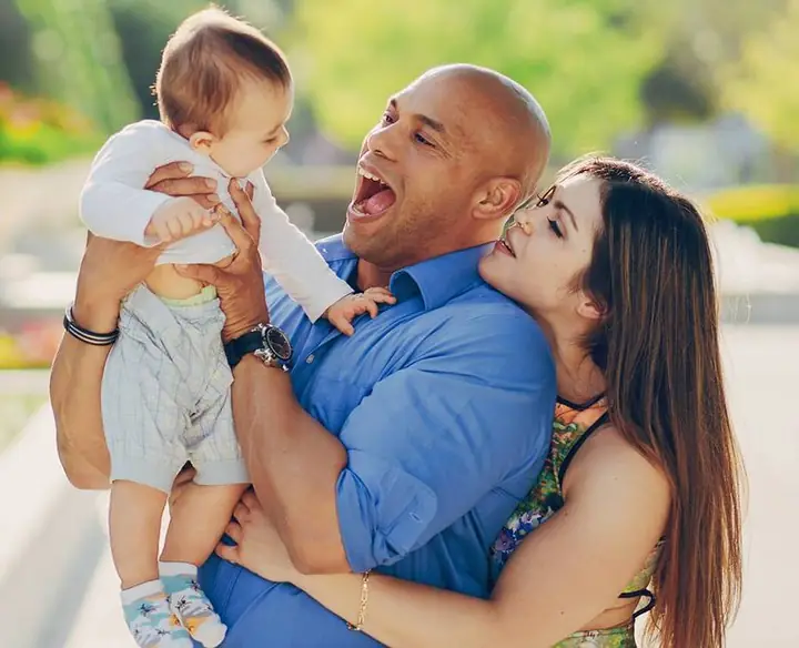 Pai sorridente levantando o bebê no colo, com a mãe abraçada a ele, num jardim